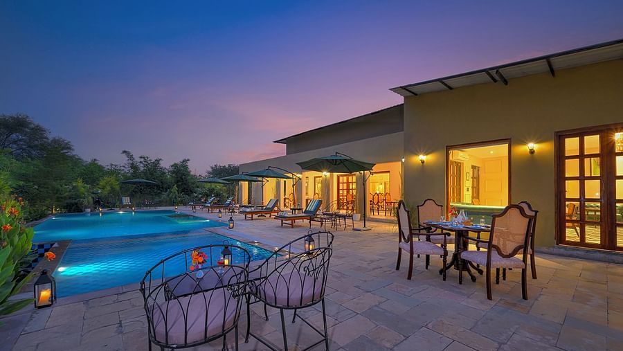 Poolside seating area with cushioned chairs and dining tables, surrounded by greenery and sun loungers under large umbrellas, illuminated by lanterns at dusk - Singhvilas, Ranthambore