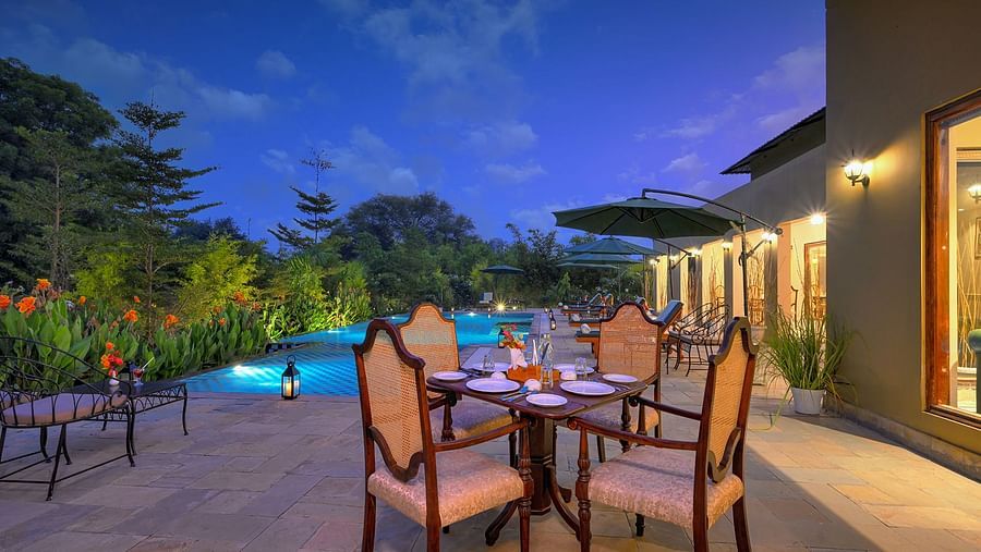 Outdoor dining setup with a wooden table and cushioned chairs near a swimming pool, surrounded by greenery, lounge chairs, and large green umbrellas under a twilight sky - Singhvilas, Ranthambore