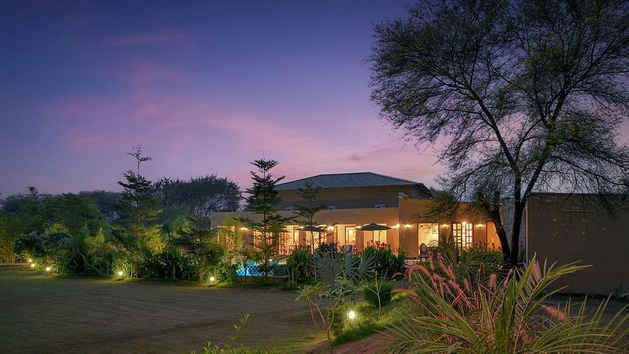 A villa with a sloping roof, warm exterior lighting, and large windows sits amidst lush greenery. A swimming pool and outdoor seating area are visible, with a twilight sky in the background - Singhvilas, Ranthambore
