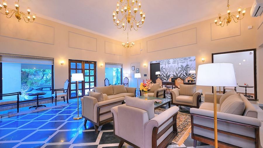 Bright hotel lounge with large windows, golden chandeliers, beige sofas, patterned black-and-white flooring, and a cosy seating area next to a wall adorned with tropical-themed wallpaper - Singhvilas, Ranthambore