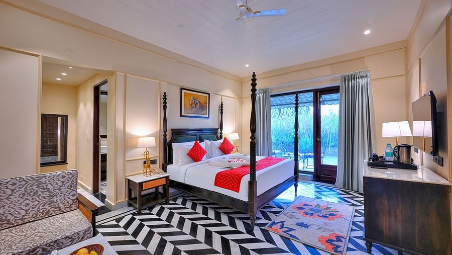 Premium Suite Cottage at Singhvilas Ranthambore features a four-poster bed with red accents, a balcony, a black and white zigzag patterned floor, comfortable seating, a TV, and tiger-themed artwork.