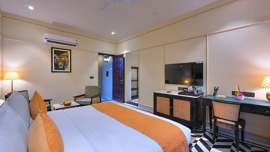 An interior view of Superior Cottage featuring a king size bed with white linens and orange accents, a television mounted on the wall, a desk with a chair, and an open doorway leading to another area - Singhvilas, Ranthambore