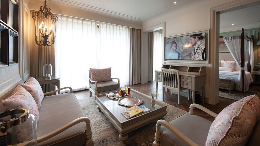 The spacious living area of a suite at The Evren, Vagator, furnished with a sofa and armchairs, a coffee table, and a mounted television. An open doorway leads into the bedroom with the four-poster bed.