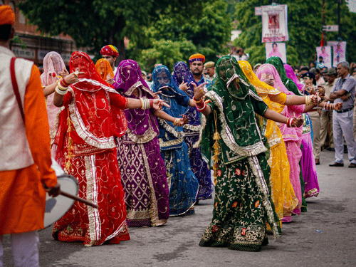 Women in embroidered saris perform traditional folk dance during Rajasthan festival procession, surrounded by drummers, spectators, and fluttering scarves outdoors