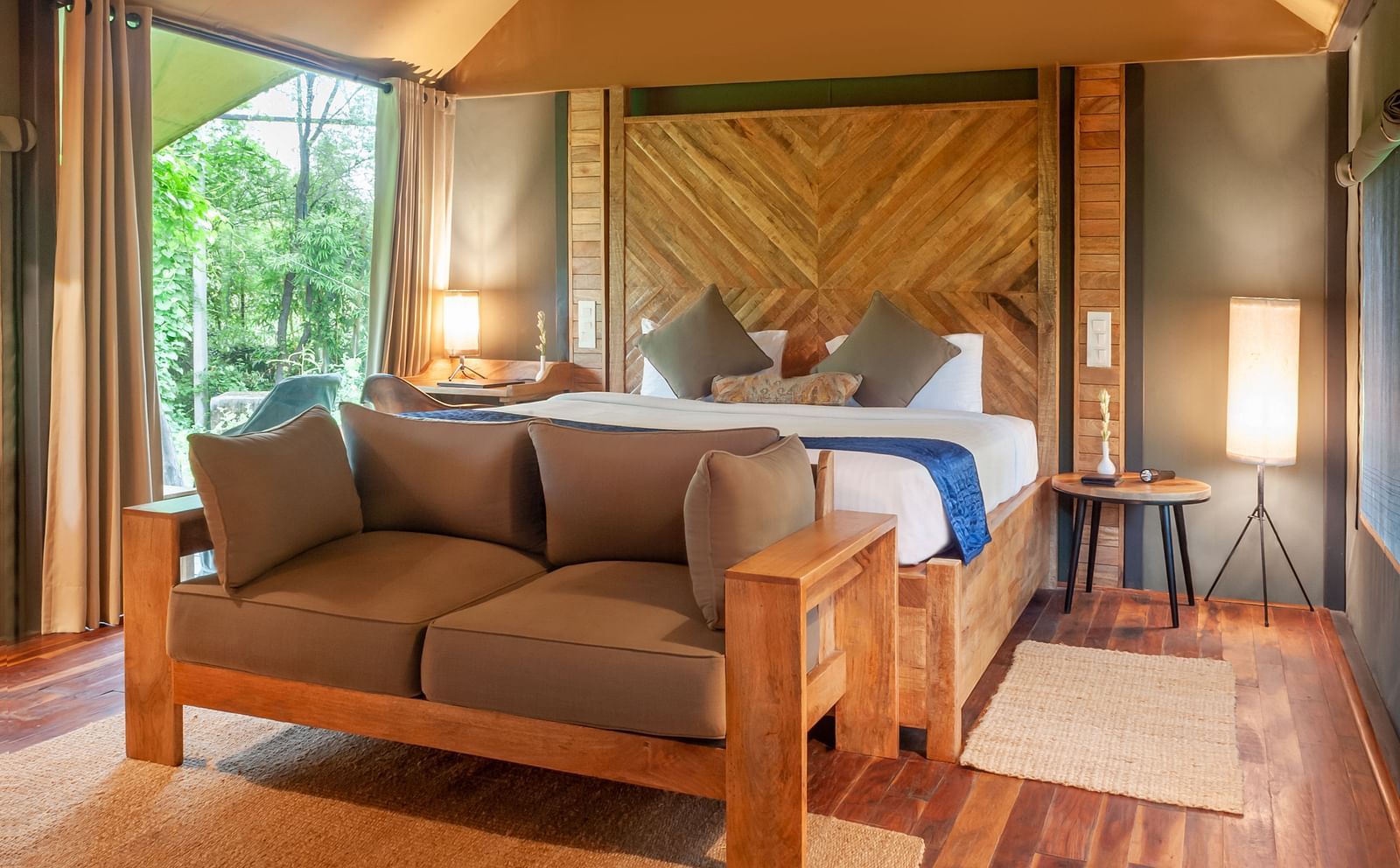 Wood-furnished room with a cushioned sofa, large bed, warm lighting, and forest views through glass windows - Trees N Tigers, Sariska