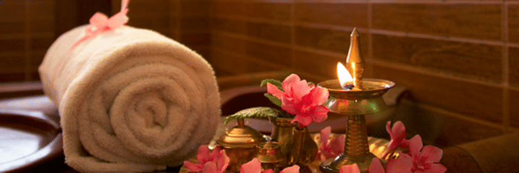A rolled white towel tied with a ribbon placed beside a decorative plate of pink flowers and a lit brass oil lamp at Ayur Ashram, Uday Samudra Leisure Beach Hotel