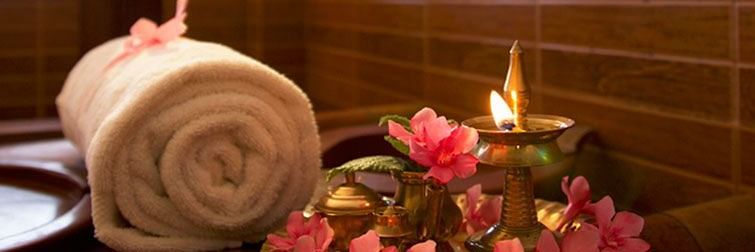 A rolled white towel tied with a ribbon placed beside a decorative plate of pink flowers and a lit brass oil lamp at Ayur Ashram, Uday Samudra Leisure Beach Hotel