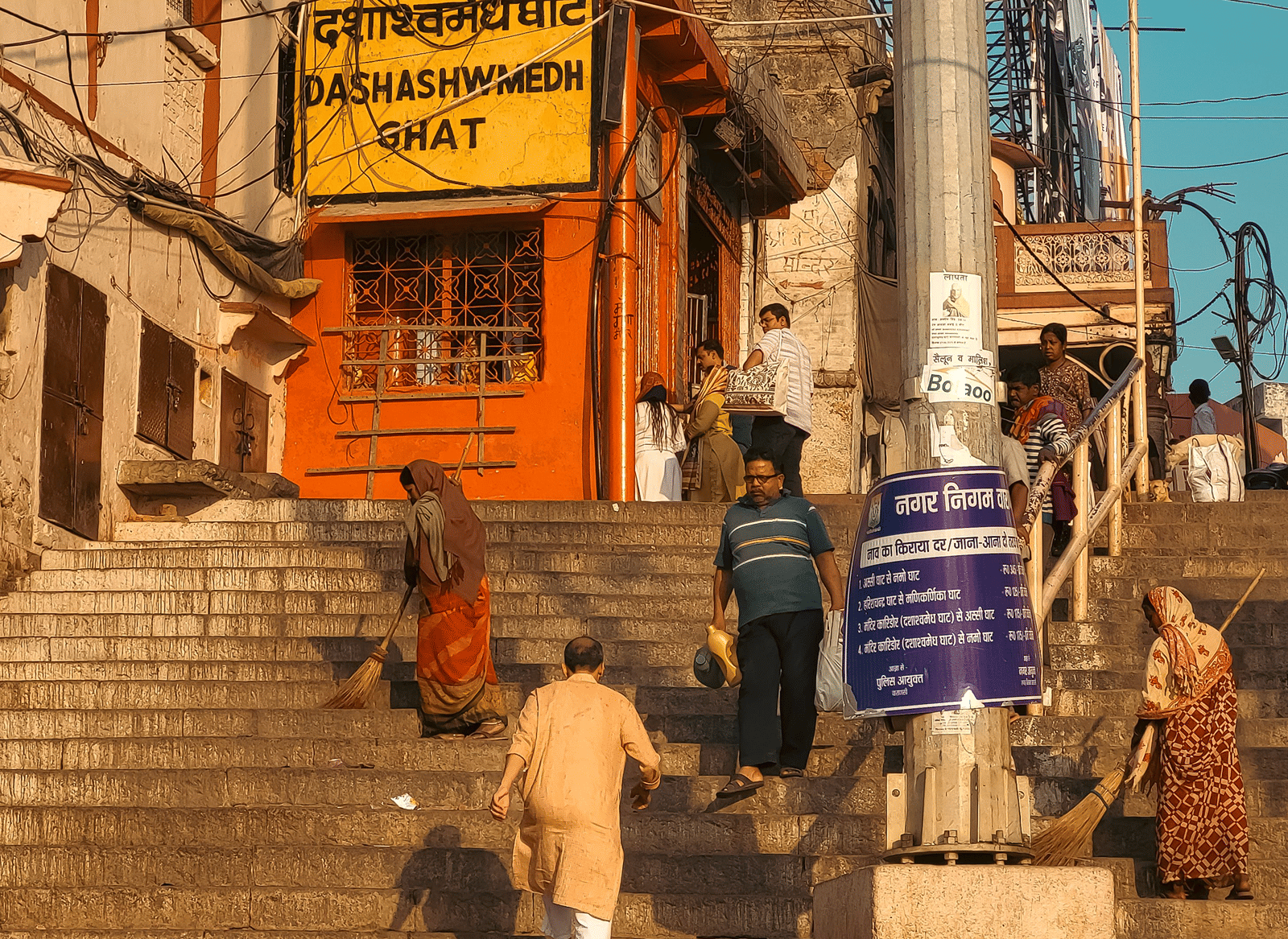 Stone steps of Dashashwamedh Ghat in Varanasi with a yellow name board on an orange building, pilgrims climbing, and 2 sweepers at work in morning light.