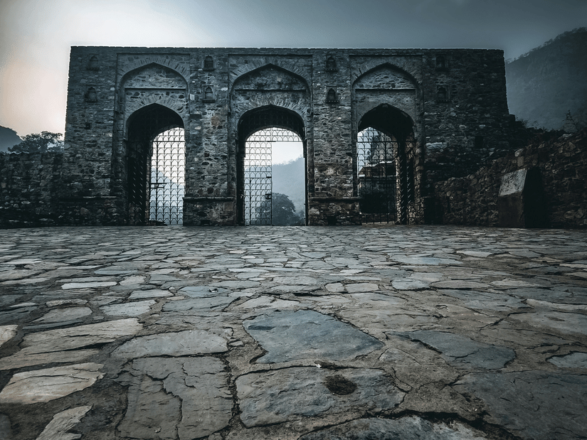 A view of the arch of the Bhangarh Fort with a stone courtyard in front of it. The Bhangarh Fort history is filled with folklore about curses.
