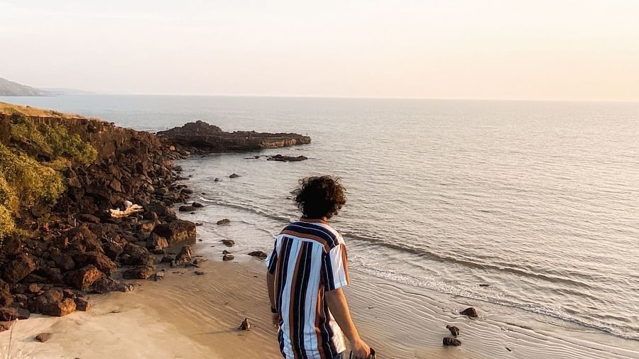  A person wearing a striped top and jeans stands on a cliff overlooking a beach and the ocean at sunrise or sunset. They are looking out at the view, and a camera or binoculars hangs around their neck.
