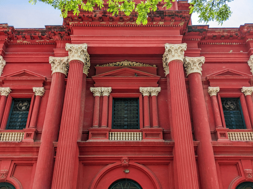 A grand crimson heritage building boasting stately columns, framed gracefully by lush green tree leaves above