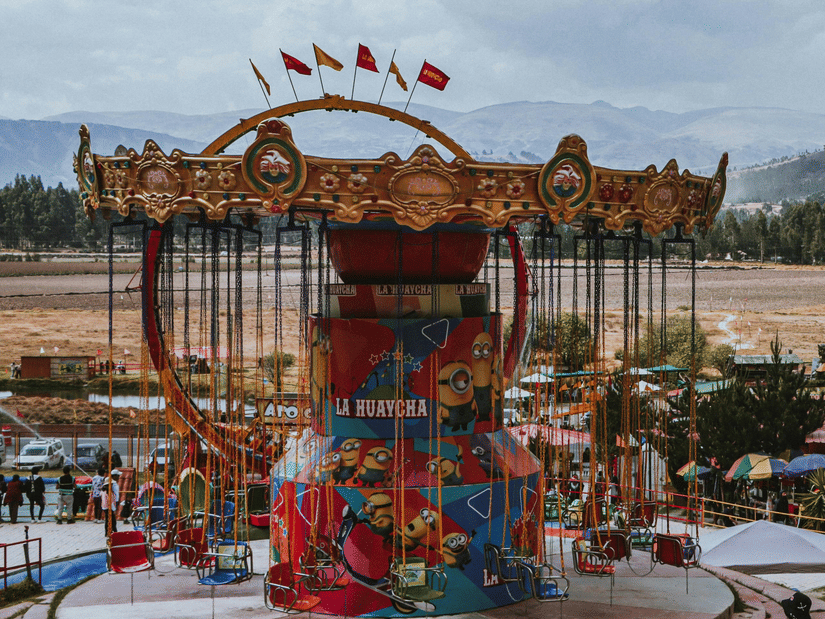 A carousel ride at an amusement park with people walking behind it, a barren land and trees in the background