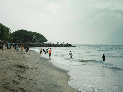 Visitors stroll along a broad sandy beach where gentle waves meet the shore beneath pale skies and a distant tree-lined coast