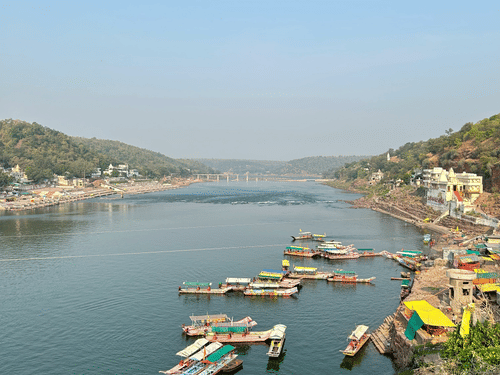 A view of the Narmada River with numerous boats on it, buildings on either side and a bridge seen in the distance. It is one of the best Bharuch tourist places to visit.