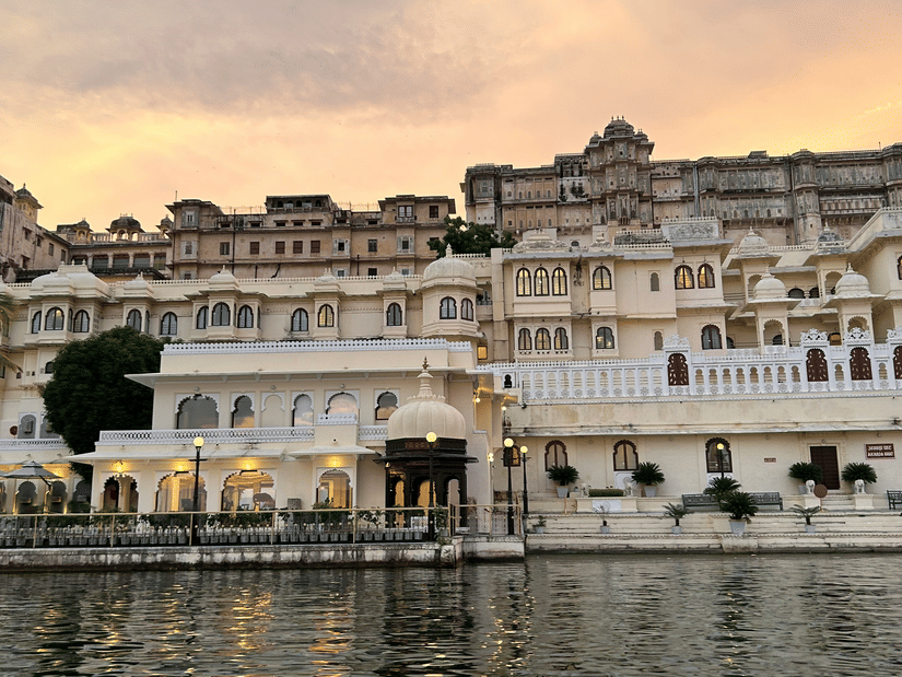 A view of the City Palace with a waterbody in front of it and different hues in the sky during sunset at Ambrai Ghat, Udaipur
