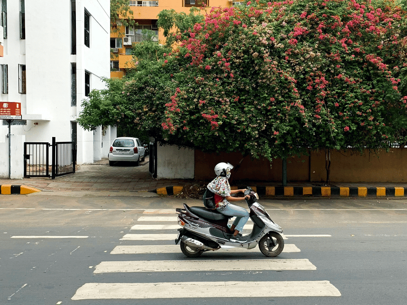 A woman riding a moped on an empty road with a tree and buildings in the background.