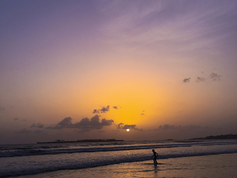 A person stands in the surf at dusk as the sun sets over the ocean, casting a golden glow across the purple sky. Aksa Beach is one of the best sunset points in Mumbai.