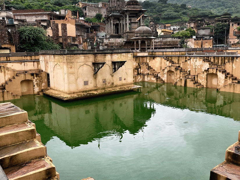 A scenic view of a traditional stone stepwell filled with green water, set against hills and old temples.
