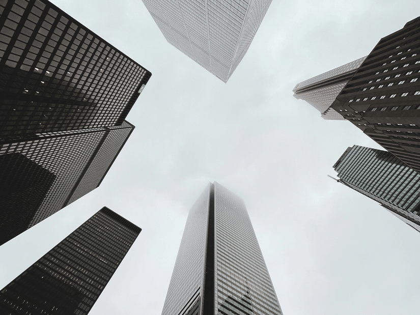 A low-angle, upward view looking straight up at several tall skyscrapers towering into an overcast grey sky.