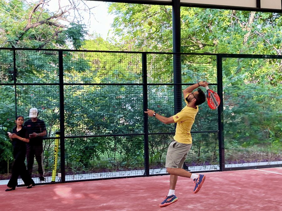 A man in a yellow shirt jumping to hit a ball on a red padel tennis court surrounded by greenery - Karma Lakelands