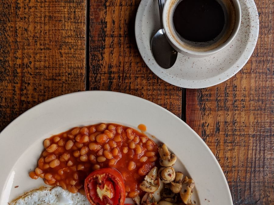An overhead shot of English breakfast in a white plate sided with a coffee served in a white mug and saucer