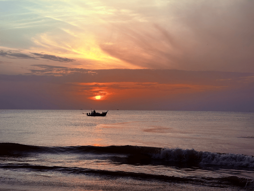 Peaceful sunset view over the ocean with gentle waves washing onto the beach and a silhouette of a boat on the horizon.