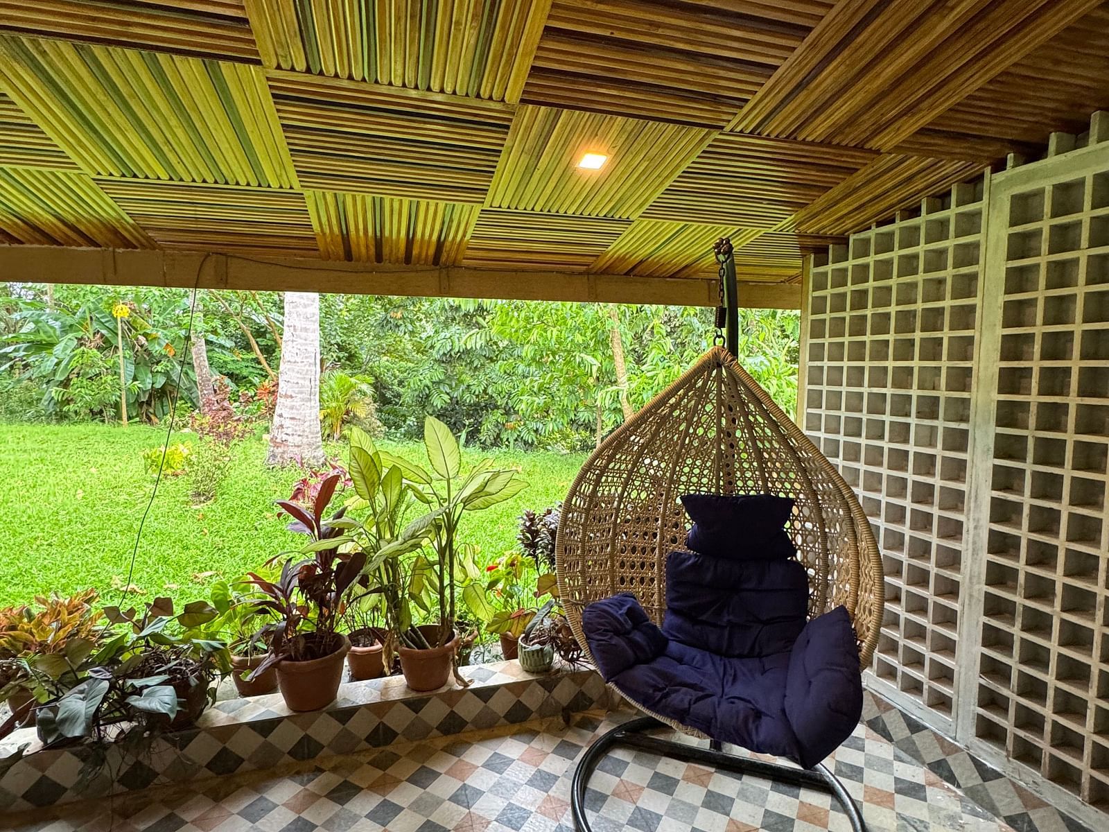A cozy outdoor seating area with a hanging wicker chair, patterned tile floor, and a distinctive wooden ceiling.