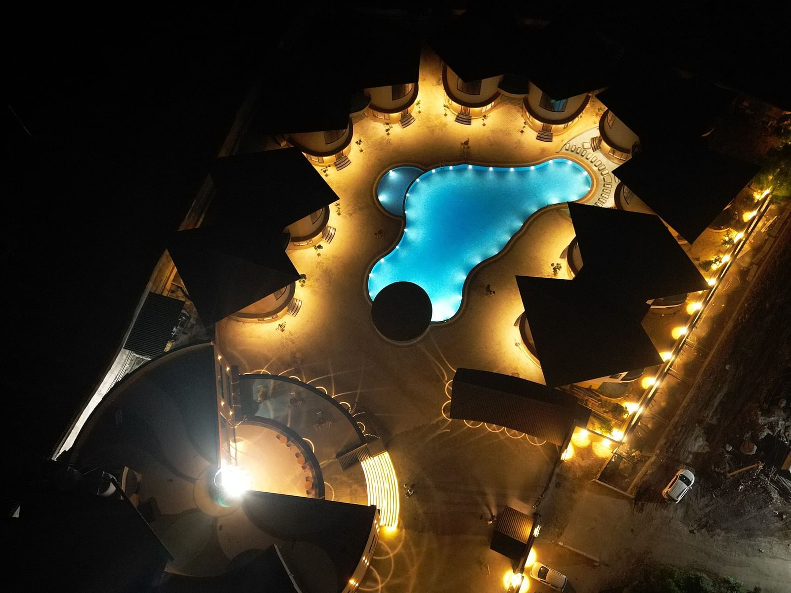 An aerial night view of The Qualia Resort Club & Brewery, Udaipur capturing a pool area, illuminated with warm lights and surrounded by lush greenery.