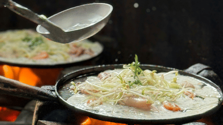 Food being cooked in a pan over open flame, with vegetables and utensils visible above the cooking station.