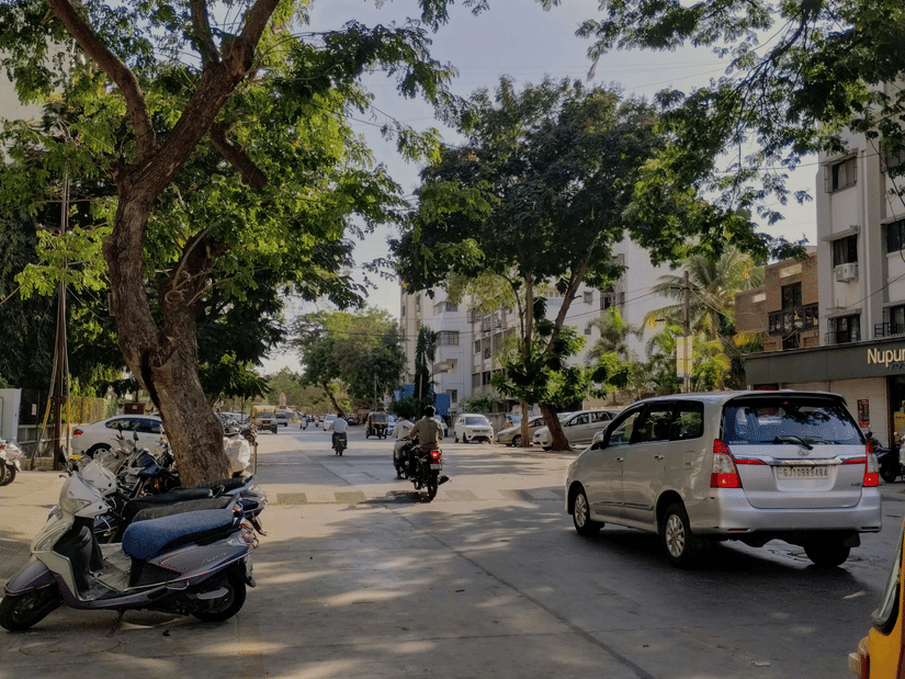 An Jamnagar street with trees, parked vehicles, and people walking along the roadside.
