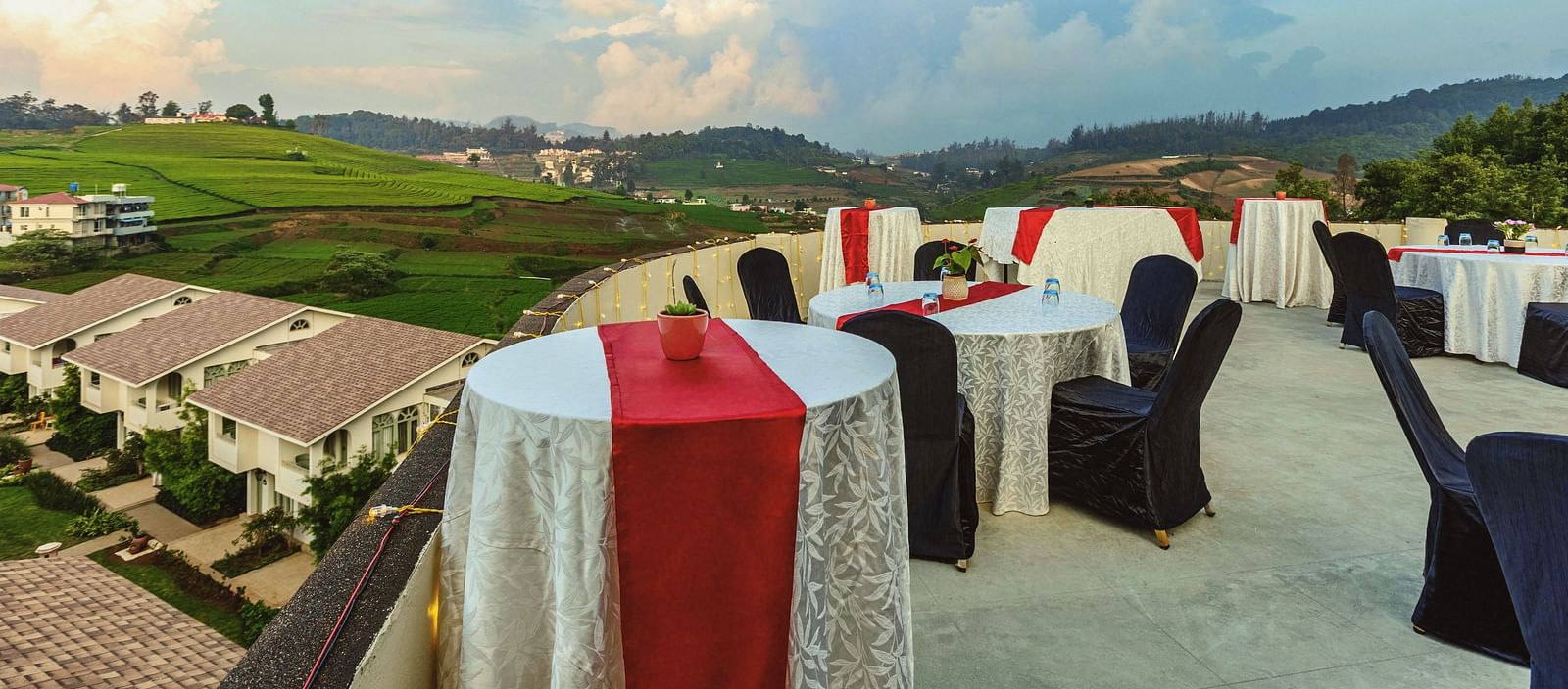 Scenic rooftop dining and event area at Mango Hill Shola, Ooty, showcasing neatly arranged tables with red and white covers, black chairs, and panoramic views of rolling hills, terraced fields, and a clear sky.