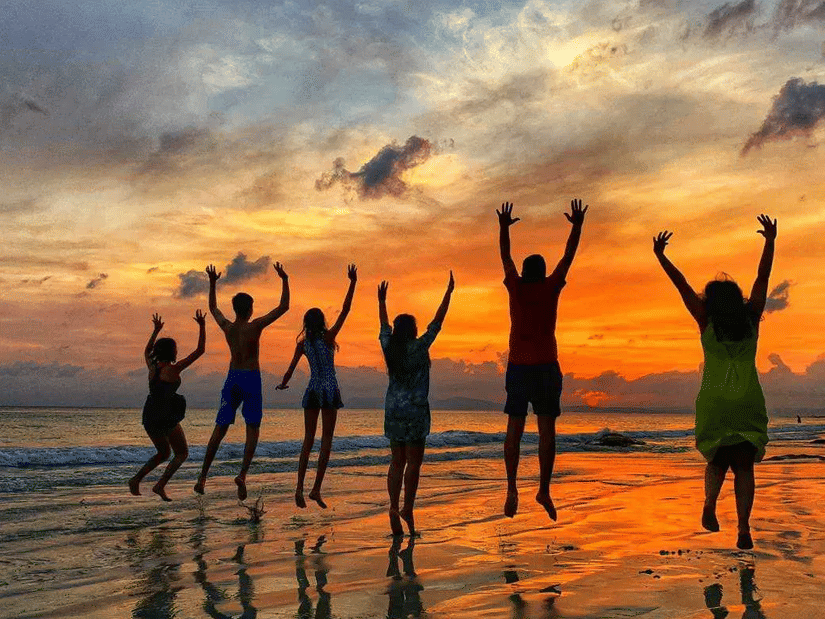A family standing on a beach at sunset, jumping with their hands raised, with their reflections visible on the wet sand and the sky filled with clouds.