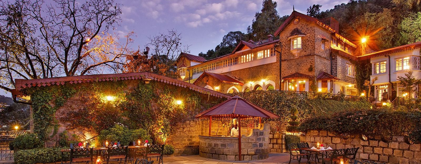 A beautiful outdoor dining area at the naini retreat, nainital. There are tables and chairs set up on a stone patio, with a fireplace in the center. The building is lit up at dusk, creating a warm atmosphere. 
