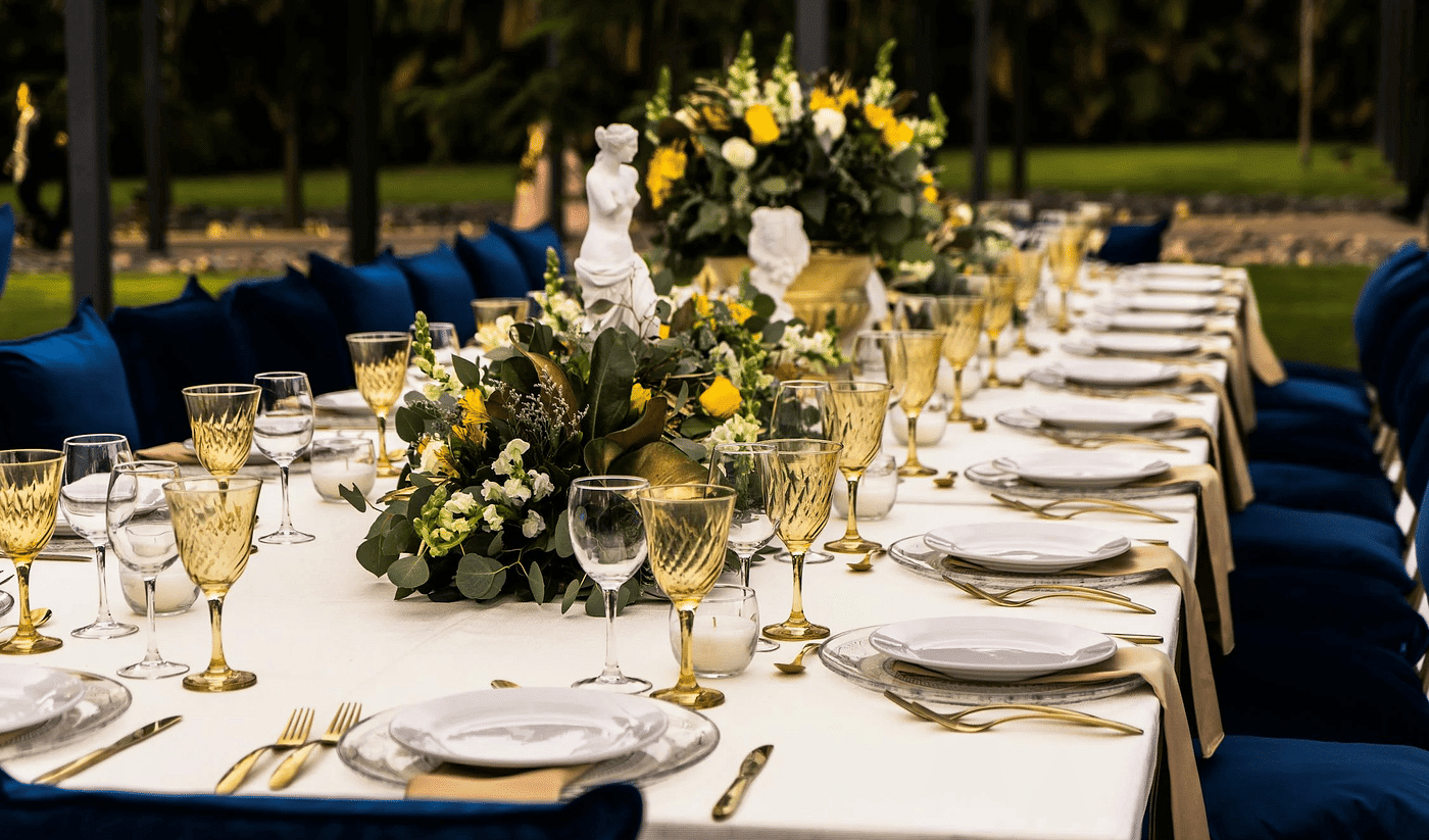 A rectangular table in an outdoor setting with cutleries arranged on it.