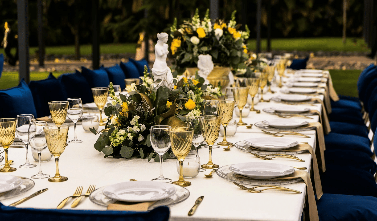 A rectangular table in an outdoor setting with cutleries arranged on it.