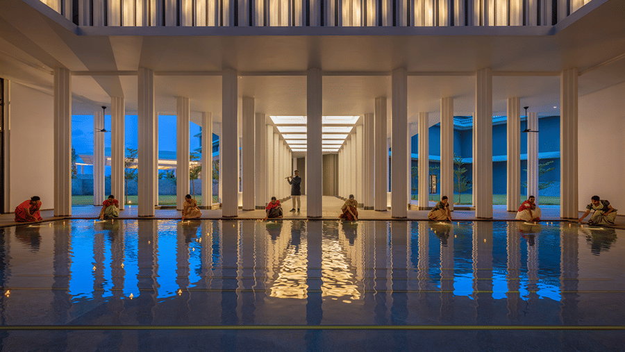 Colonnaded courtyard with a reflecting pool at dusk, women in traditional dress kneeling at the edge and a flautist at The Residency Signature Madurai.