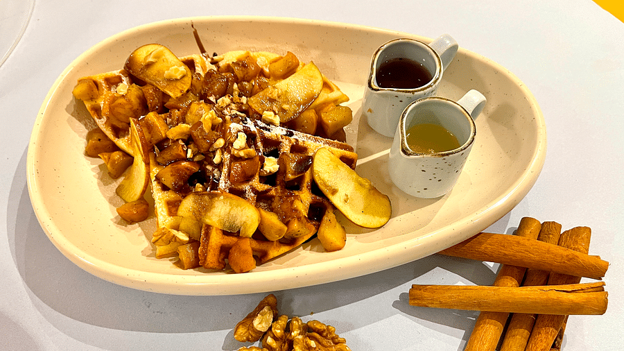An image of a nutty waffle topped with fruits and chocolate, served on an oval plate, with walnuts and cinnamon placed on the table.