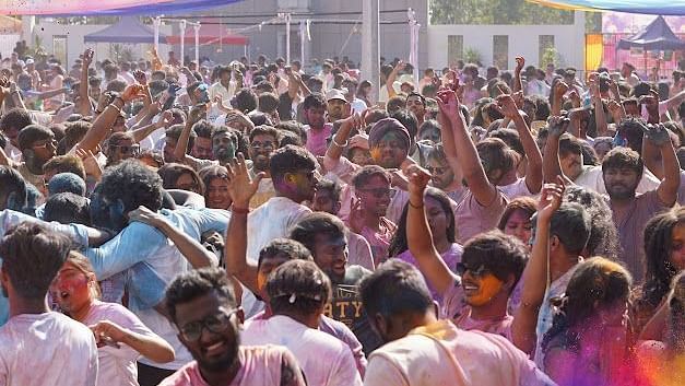 Massive crowd enjoying Holi festival, drenched in colours, raising hands, and dancing joyfully.