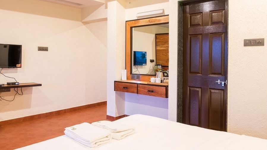 A room with a white bed, a dark wood door, a mirror above a wooden dresser, and a mounted TV.