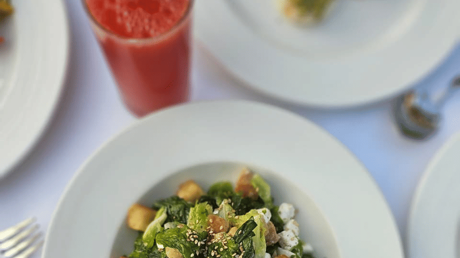 A mixed greens salad with croutons and shredded cheese placed neatly on a plate beside a glass of juice on a clothed table at A.S. Hotels, Khajuraho.