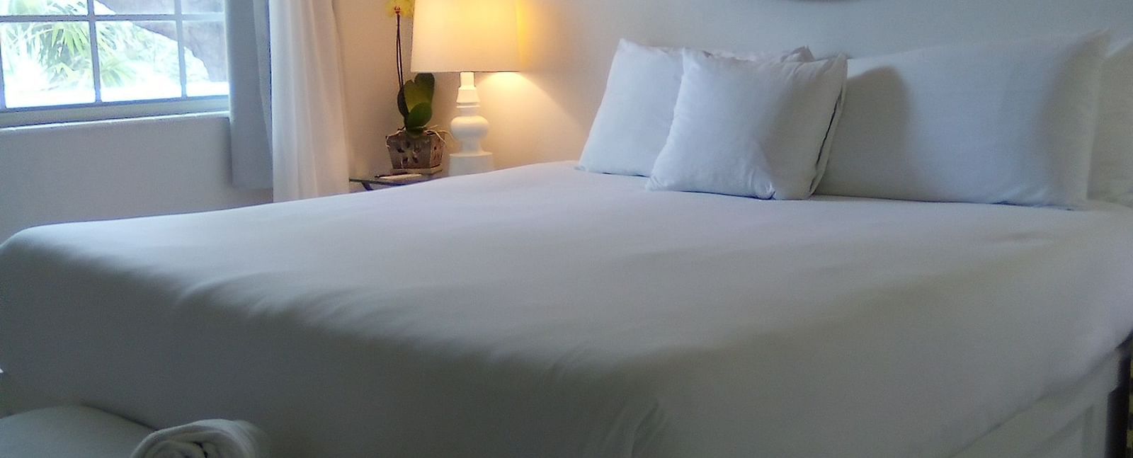 Bed at Casa Morada with white linens, towels on a bench at the foot, and a window with curtains in the background.