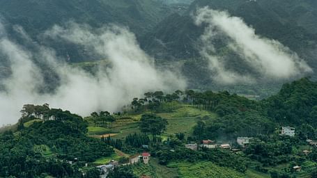 An aerial view of a village next to a mountain with mist flowing into the village