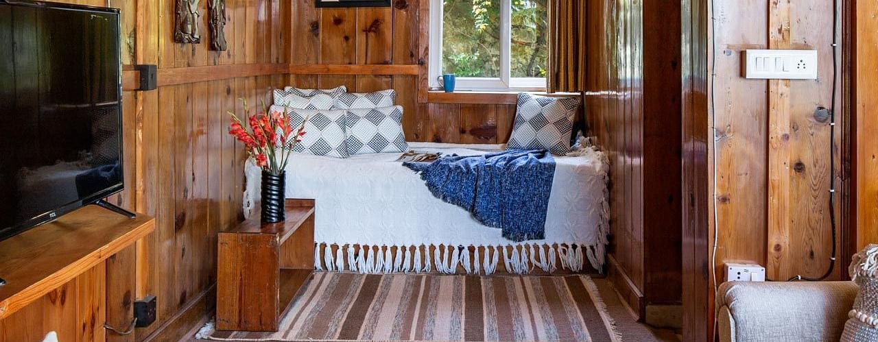 An image of a wooden bedroom with single bed, bedside table, an open window, a sofa, and flat TV screen - Avalon Cottages, Kanatal
