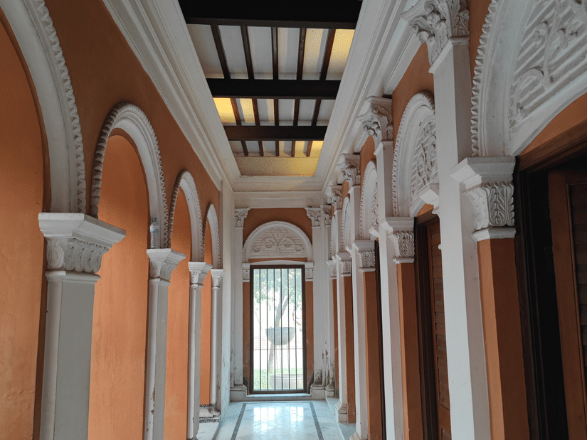 Long heritage corridor with graceful arches, polished flooring, and wooden doors, reflecting the elegant design of a traditional Chettinad mansion.