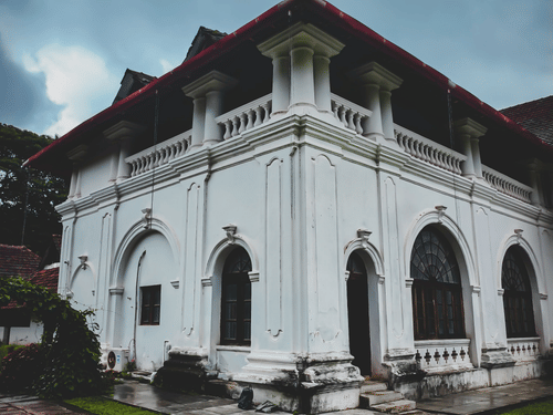 A historic white heritage building features ornate arches, columns, and a red-tiled roof amid lush greenery.