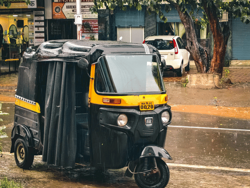 An auto rickshaw parked on roadside near trees and buildings, with wet road surface visible in the foreground.