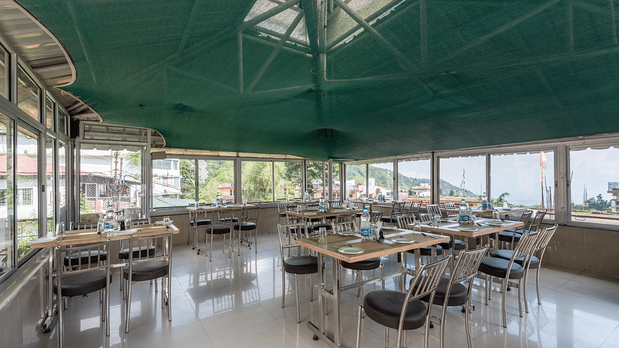 A coffee house with tables and chairs under a green canopy, featuring large windows offering an outdoor view. The tables are set with plates and bottles of water - DLS Meghavan Resort, Dharamshala