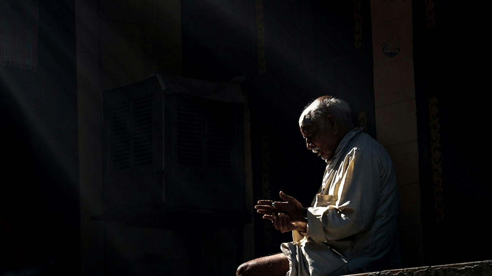The image shows an elderly man sitting on a ledge, possibly in a dimly lit or outdoor setting. The man is positioned to the right of the frame, with his hands clasped together in what appears to be a contemplative or prayerful gesture. Sunlight streams in 