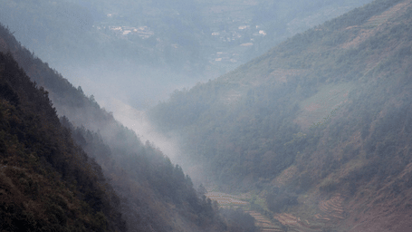 Misty mountain valley at dawn with layered ridges, terraced fields and a small village nestled between forested slopes.
