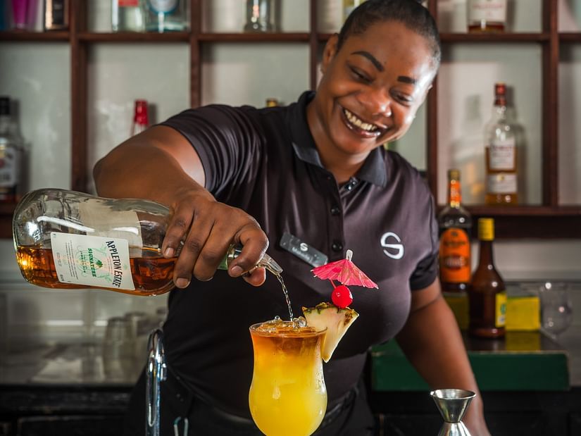 A bartender pouring a cocktail into a glass containing  a fruit peel and a cocktail umbrella - S Hotel Montego Bay, Jamaica